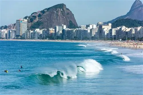 Волны разбиваются о берег оживленного пляжа Копакабана в Рио-де-Жанейро в солнечный день. Люди купаются в океане.