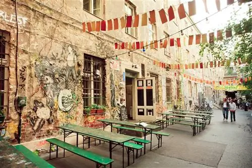 Внешний вид модного бара в Берлине, расписанный граффити. Снаружи стоят простые зеленые столы и скамейки, а над головой висит флаг.