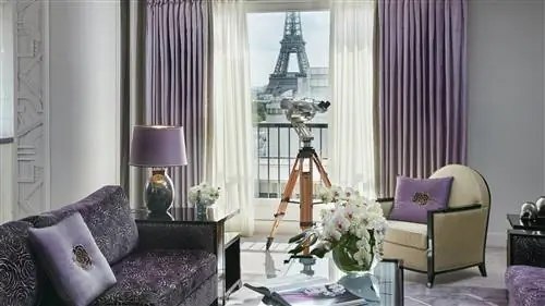 Люкс Eiffel в парижском отеле Plaza Athenee украшен роскошным фиолетовым диваном, лавандовыми шторами, серой лампой в форме яйца с лавандовым абажуром, кремовым креслом с лавандовой декоративной подушкой и журнальным столиком с большими белыми цветами в зеленом цвете. ваза. Через окно вид на нижнюю часть Эйфелевой башни с телескопом, расположенным для облегчения обзора.