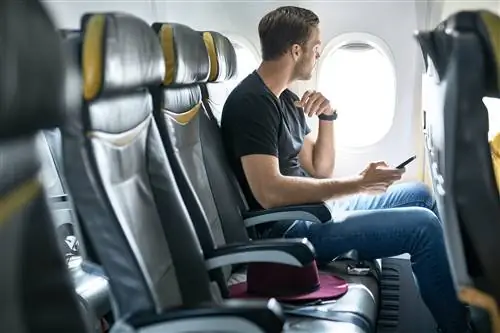 Пассажир мужского пола сидит в самолете рядом с окном с мобильным телефоном.