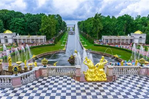 Клетчатая черно-белая черепичная терраса с золотыми статуями и фонтанами за ней. Канал ведет к реке, и можно увидеть толпы людей, слоняющихся вокруг водных объектов.