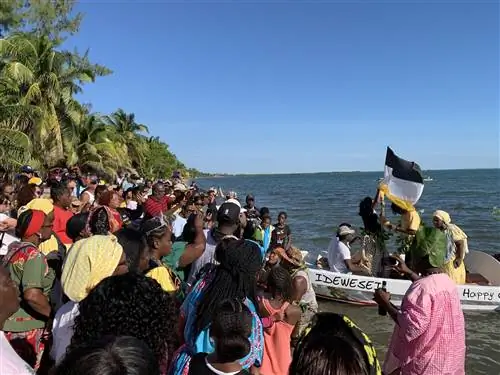 Большая группа людей стоит на берегу, наблюдая, как пара лодок, наполненных актерами Гарифуна, воссоздает День поселения Гарифуна.