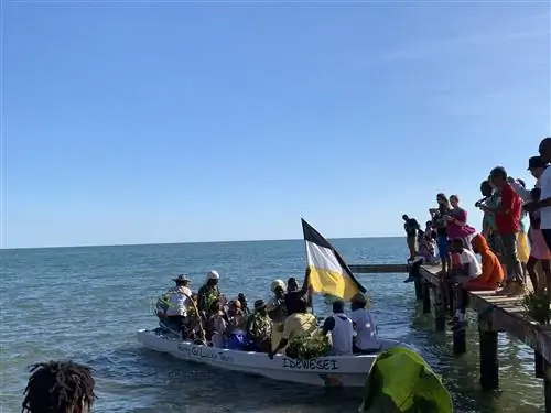 Большая лодка, наполненная актерами, воссоздает День поселения Гарифуна, а группа людей наблюдает за ним с деревянного пирса.