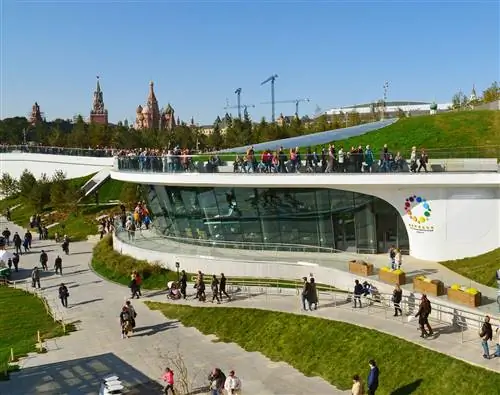 Зарядье - ландшафтно-архитектурный парк. Многие люди прогуливаются по бетонным дорожкам, окружающим округлое здание в парковой зоне.