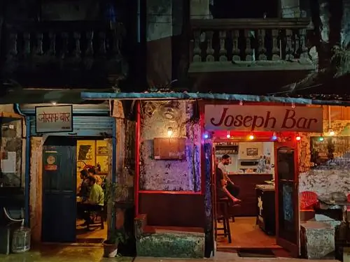 Тускло освещенный фасад бара Joseph в Гоа подсвечен красными и фиолетовыми гирляндами и теплым светом, льющимся из самого бара. Вход находится под темным балконом, а внутри несколько посетителей заполняют дверные проемы.