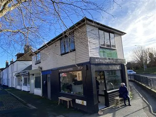 Человек в шапке и теплом пальто сидит на скамейке возле кафе Mit Milch в ясный день.