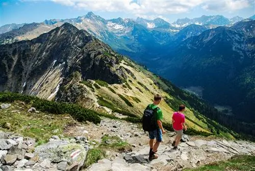 Двое туристов, одетых в летнюю одежду, идут по каменистой тропе вниз по склону холма. Вдали простирается горный хребет