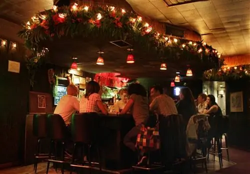 Шесть посетителей сидят вокруг бара в Tonga Hut в Лос-Анджелесе, где красные тропические цветы переплетаются в гирлянде из пальмовых листьев, усеянной огнями над барными стульями в стиле середины века.
