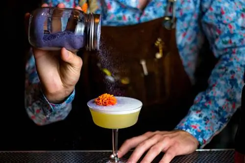 Бармен посыпает пенный коктейль фиолетовым порошком для украшения с цветком апельсина наверху.