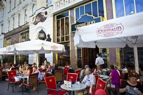 Группа людей пьет кофе за маленькими столиками под большими белыми зонтиками рядом с кафе Gerbeaud.