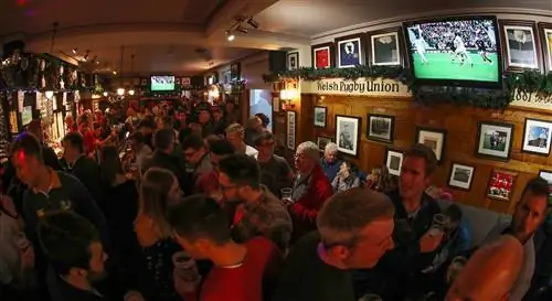 Вид на интерьер пабов City Arms в Кардиффе перед матчем по регби в городе. Паб битком набит людьми, большинство из которых одеты в красные футболки сборной Уэльса по регби.