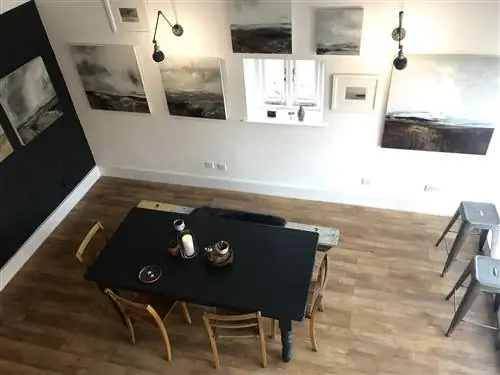 Вид вниз с антресольного этажа в комнате в Gallery at Home, Уск. Черно-белые пейзажи висят на монохромных стенах, а минималистичный обеденный стол и стулья стоят в центре комнаты.