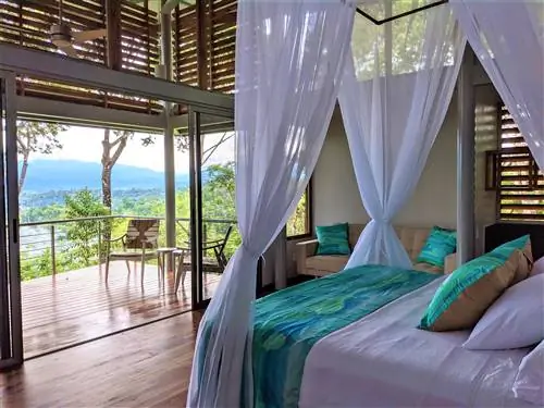 Внутренний снимок внутренней части Cielo Lodge. Здесь есть кровать с белыми занавесками, ниспадающими с каркаса кровати, и диван с подушками ярко-зеленого цвета. С балкона открывается вид на пышную загородную местность.