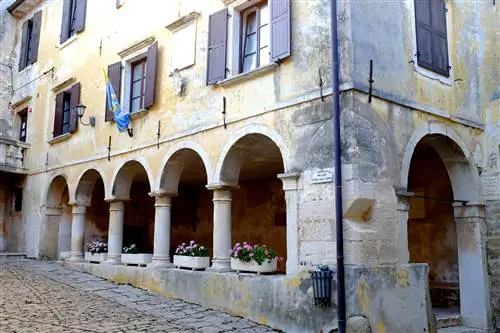 Обветренный серо-желтый экстерьер одной из лоджий в Грожняне на границе с Хорватией украшен оконными ящиками с розовыми цветами в арках чуть выше уровня улицы, окнами с коричневыми ставнями и сине-желтым флагом, обмотанным вокруг шеста.