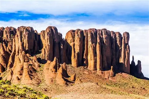 Луковицеобразные, почти пальцевидные образования красных скал поднимаются со дна долины; долина усеяна зеленой кустарниковой растительностью.