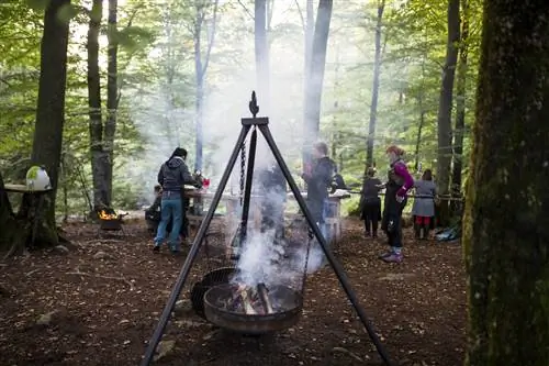 Группа из восьми-десяти человек собирается вокруг деревянных столов в лесной роще, держит напитки и наблюдает, как поднимается дым от большого металлического кухонного штатива в центре кадра. К треноге на трех цепях подвешена металлическая жаровня с горящими поленьями. Вдалеке ярко горит еще одна приподнятая яма для костра.