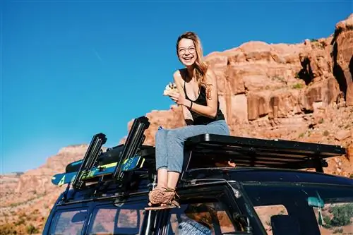 Katie_Boué сидит на багажнике на крыше своего фургона в серых спортивных штанах и черной майке, улыбаясь, с волосами, собранными в конский хвост, и круглыми очками на лице. Она держит кусок пиццы.