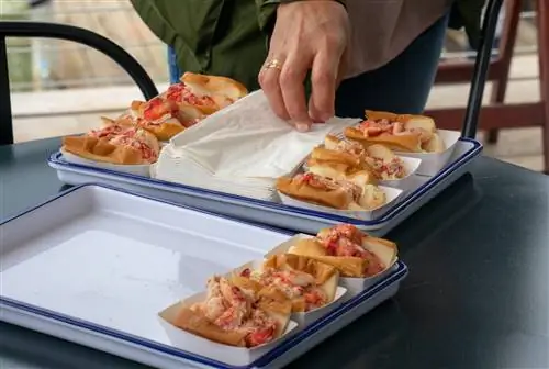 Рука с золотым обручальным кольцом тянется за салфеткой из стопки между двумя рядами роллов с лобстерами штата Мэн в ресторане Luke's в Портленде. Роллы находятся в маленьких бумажных лотках и заполнены ярко-красным и розовым мясом омара в белых и синих эмалированных лотках.