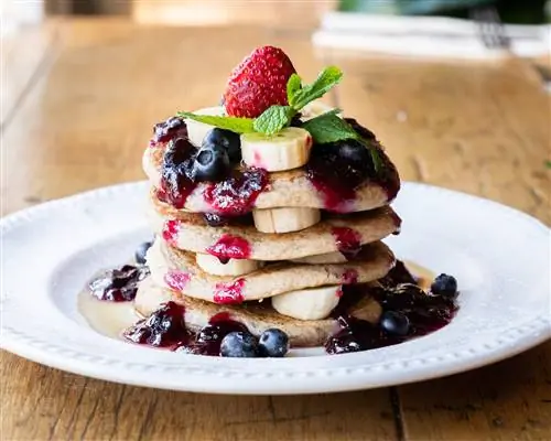 Огромная стопка веганских блинов в кафе «Миранда» с бананами, черникой и красным компотом на белой тарелке, посыпанной сахарной пудрой.