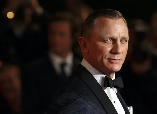 Актер Джеймса Бонда Дэниел Крейг одет в смокинг и смотрит в камеру; фон темный и в мягком фокусе.