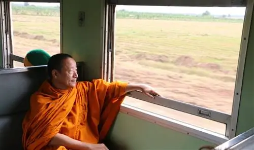 Монах смотрит в окно на поезд в Таиланде, проезжающий мимо зеленой сельской местности.