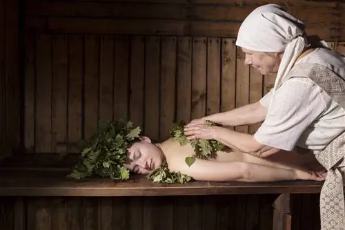 Обнаженная женщина лежит лицом вниз на деревянной скамье. Другая женщина в кремовой одежде кладет себе на спину кучу листьев.