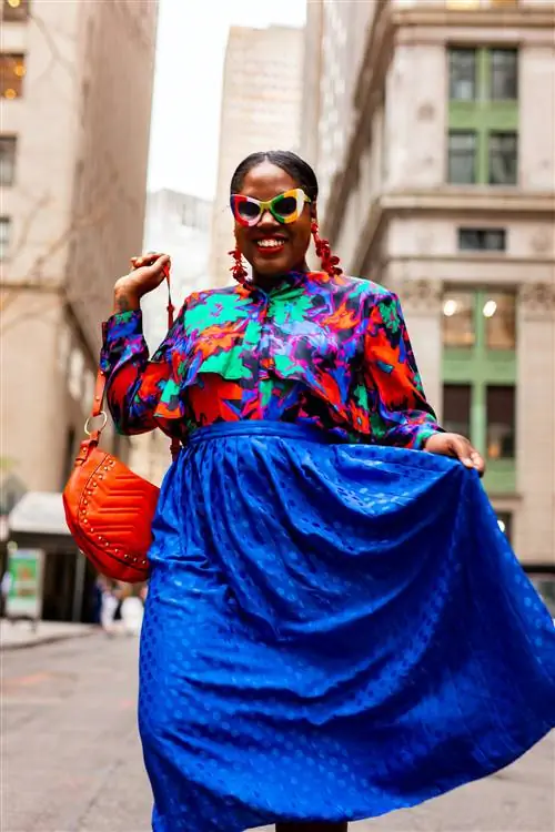 Улыбающаяся женщина в цветочной блузке и синей юбке улыбается, неся красную сумочку