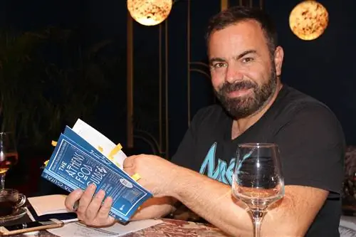 Василис Каллидис уверенно улыбается в камеру, держа экземпляр своей новой книги «Афинский гастрономический справочник» под теплым светом ламп из дутого стекла, свисающих с потолка ресторана. Он одет в черную футболку, и перед ним стоит бокал с вином. У него короткая борода и темные, коротко подстриженные волосы.
