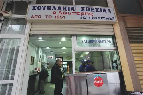 Пожилой джентльмен в черных брюках и черном пальто ждет у прилавка стойки Lefteris suvlaki в Афинах, Греция, в то время как двое сотрудников в синих рубашках готовят еду на кухне из нержавеющей стали.