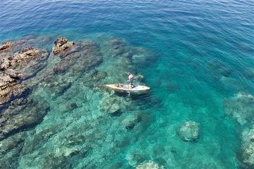 Снято сверху: мужчина гребет на SUP вдоль скалистого берега по бирюзовой воде.