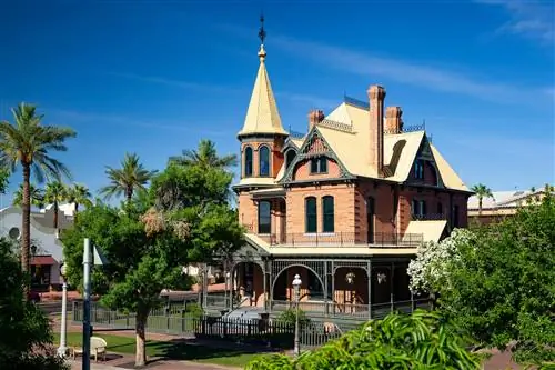 Башни и желтая крыша дома-музея Россона в викторианском стиле в парке Херитедж-сквер, Феникс, Аризона, США, выглядывают из-за зеленых деревьев ясным днем.