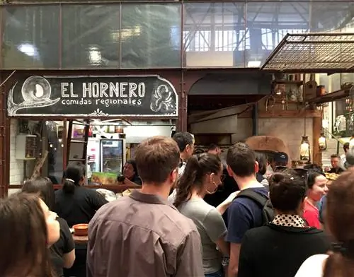 Толпа людей выстраивается в очередь за блинчиками с мясом у прилавка El Hornero на крытом рынке. На доске нарисована вывеска киоска.