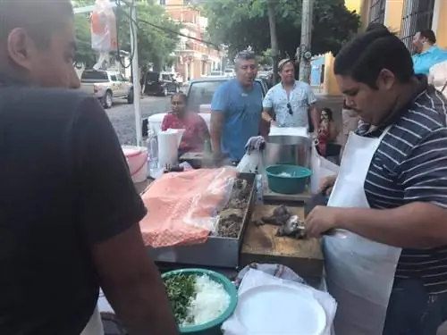 Мужчина режет мясо на деревянной разделочной доске в киоске с уличной едой в Пуэрто-Валларта. Толпа клиентов ждет, когда их обслужат.