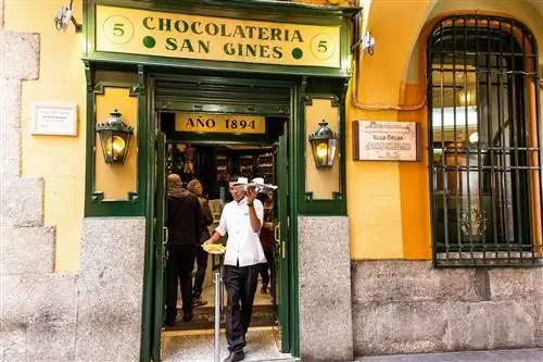Официант с тарелками чуррос и шоколада выходит из Chocolatería San Ginés Madrid; экстерьер окрашен в желтый цвет с зеленой дверной рамой и вывесками.