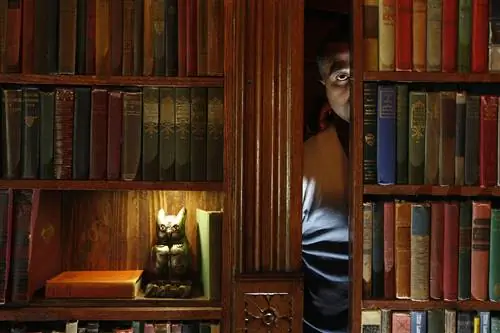 Мужчина выглядывает из-за потайной двери за книжным шкафом. На полке под ярким светом стоит золотая статуя совы.