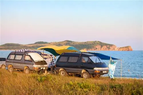 На берегу моря припаркованы два фургона Toyota с желтым и синим брезентом и складными навесами, чтобы сделать импровизированный кемпинг более комфортным.