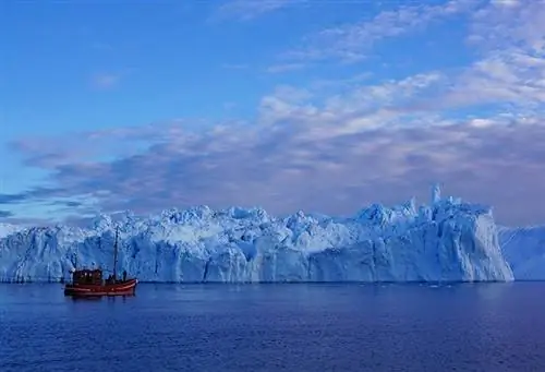 Красный парусник кажется карликом на фоне скалистого куска ледника Илулиссат, голубого обнажения в водах залива Диско, Гренландия.