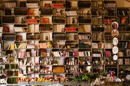 Стена с разделенными книжными полками заполнена книгами; на некоторых полках также есть винные бутылки и свежие тыквы. Стол с привлекательными фруктами и овощами стоит под книгами. Стена с разделенными книжными полками заполнена книгами; на некоторых полках также есть винные бутылки и свежие тыквы. Стол с привлекательными фруктами и овощами стоит под книгами.