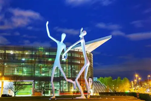 Высокие человекоподобные скульптуры стоят перед стеклянным зданием под ночным небом.