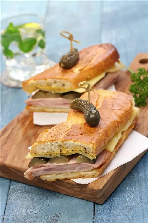 Длинный прессованный сэндвич разрезается пополам и искусно кладется на разделочную доску. Сэндвич фарширован ветчиной, сыром, свининой и солеными огурцами. Через каждую половинку бутерброда деревянной палочкой протыкают рассол.