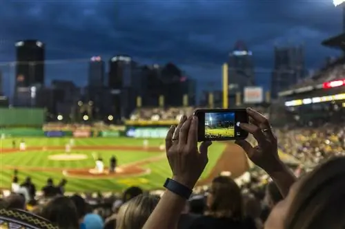 Ночью на освещенном бейсбольном стадионе женщина держит в руках мобильный телефон и делает снимок. Ее руки и телефон в фокусе, а остальная часть сцены размыта.