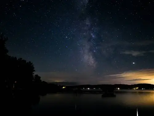 На фотографии с длинной выдержкой видны звезды и Млечный Путь, сверкающие на фоне темно-синего неба над озером Мичиган, с теплым янтарным светом нескольких домов и минимальной застройкой внизу кадра, подчеркивающей береговую линию.