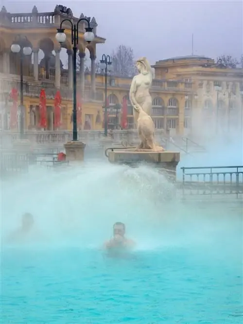 Двое мужчин стоят под форсунками в ярко-голубом бассейне, вокруг них поднимается пар. За ними характерные желтые стены курорта и мраморная статуя женщины.