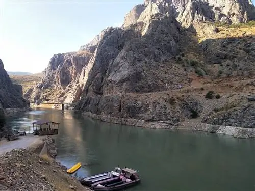 Широкая река с двумя пришвартованными лодками и небольшой деревянной хижиной, которая, кажется, висит над рекой. На противоположном берегу высоко возвышаются пыльные коричневые скалы и скалы.