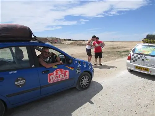 Синий европейский автомобиль припаркован рядом с белым хэтчбеком с задним стеклом, на котором напечатана карта. Между автомобилями стоят два участника «Монгольского ралли», изучающие карту. Пейзаж вокруг - голая пустыня.