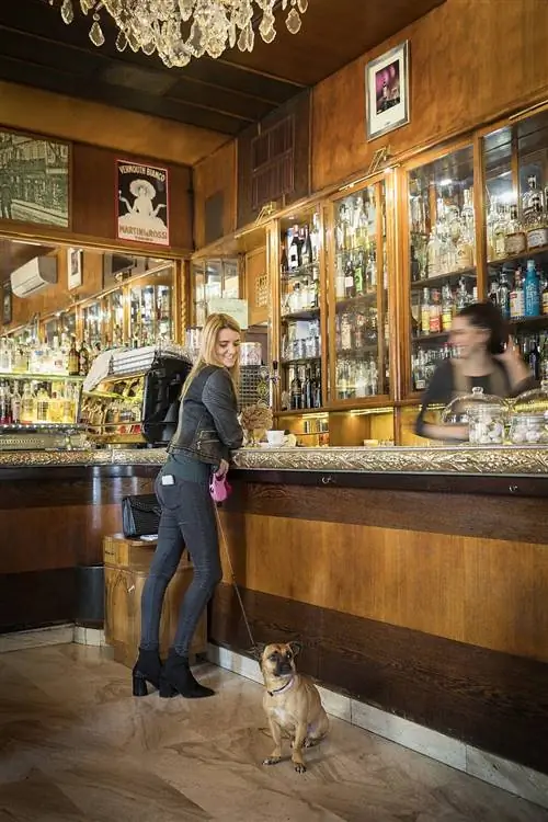 Молодая женщина стоит в традиционном баре с деревянными панелями, пьет кофе, смотрит на свою собаку и улыбается.