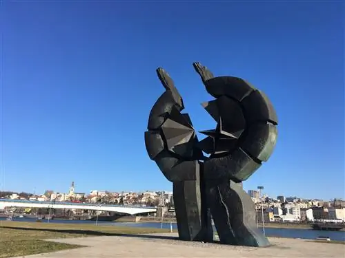 Памятник из черного камня с абстрактными руками, тянущимися вверх, образует ломаный круг. Он стоит у реки.