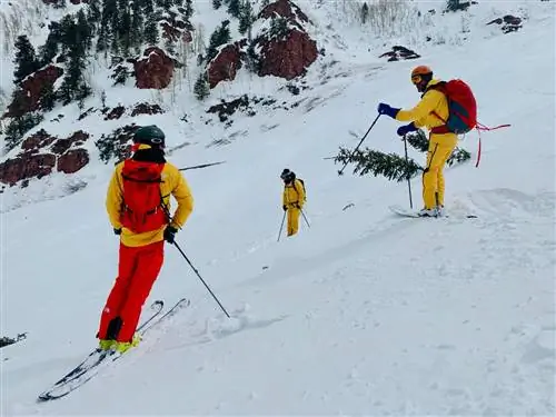 Трое лыжников спускаются по склону в Аспене, покрытому густым снегом. Все одеты в желтые и красные штаны FutureLight и куртки от North Face. На заднем плане большие валуны и деревья.