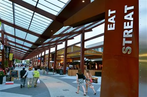 Люди идут по мощеной улице под навесом из стекла и стали. По обеим сторонам есть бары и рестораны, а на переднем плане вывеска с надписью «Eat Streat» заглавными буквами.