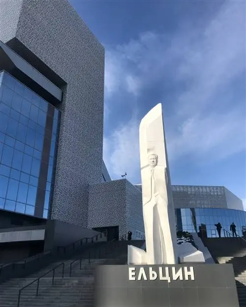 Между двумя лестничными пролетами стоит высокая белая рельефная скульптура, изображающая человека. На заднем плане большое угловатое современное серое здание.